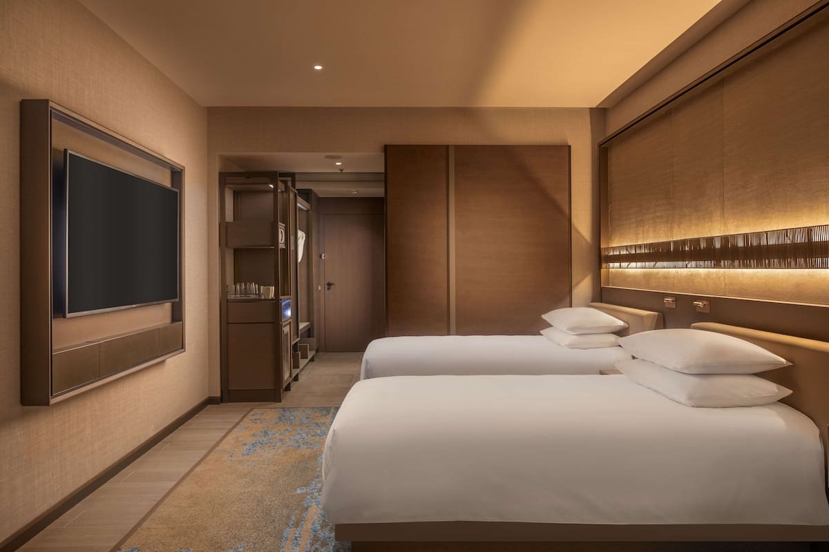 Une chambre d’hôtel dotée d’un grand téléviseur à écran plat fixé au mur, d’un dressing, d’un lit avec du linge de maison blanc et d’une tête de lit équipée d’un éclairage intégré.