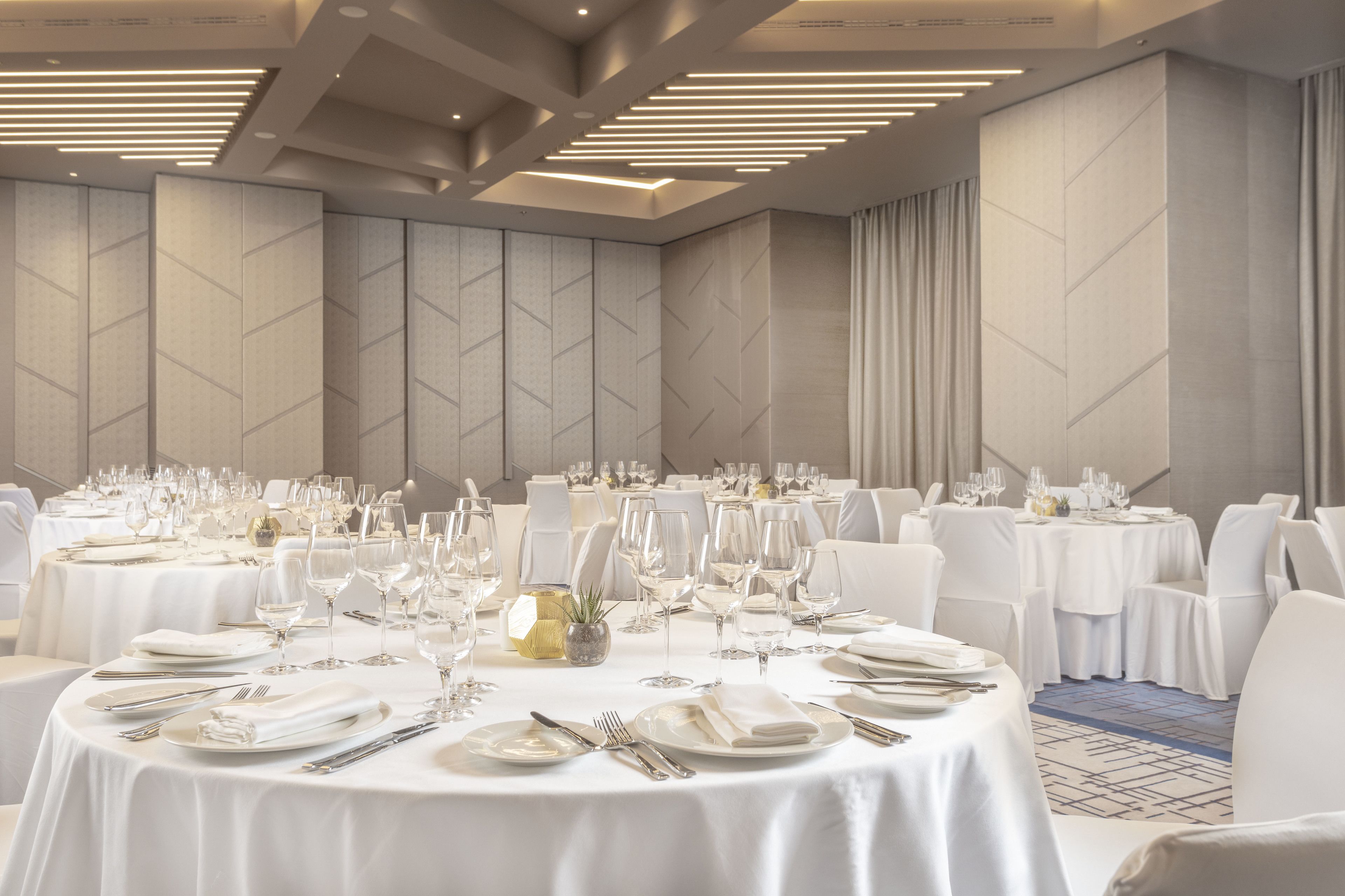 Un salón de banquetes con mesas redondas dispuestas para cenar, con manteles blancos, platos, cubiertos y copas de vino.