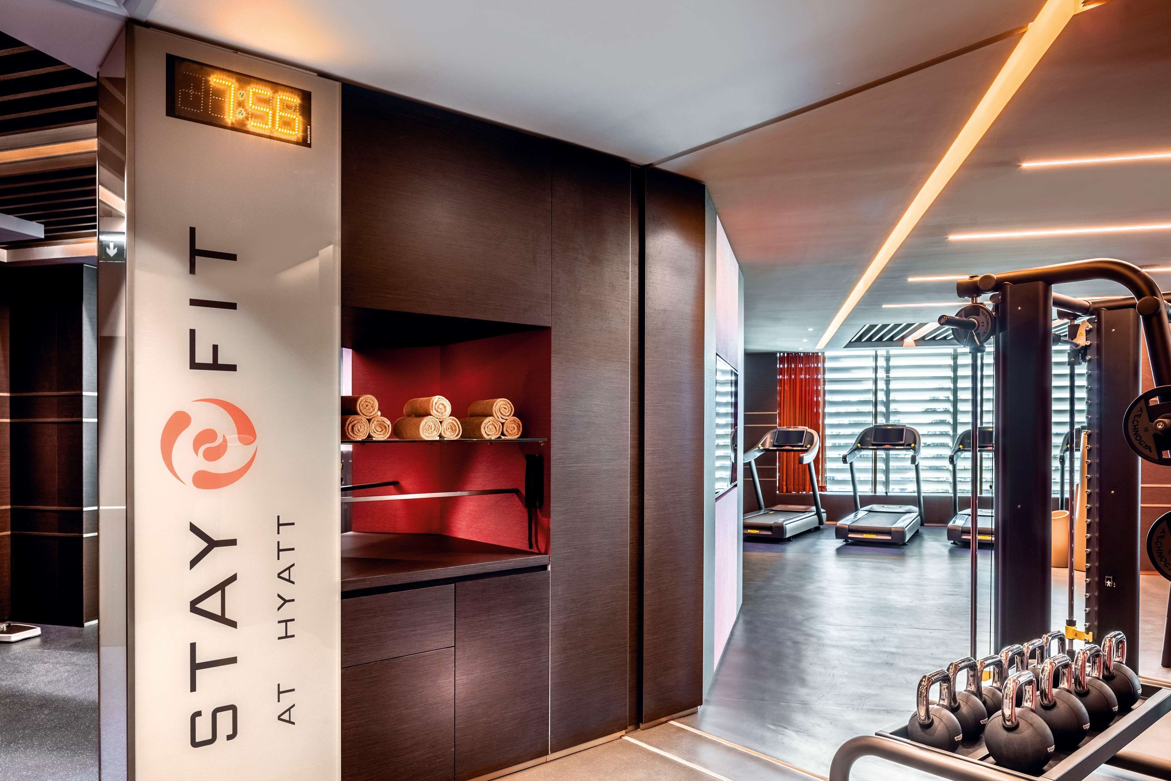 Un gimnasio moderno en un hotel, con reloj digital, estantes para pesas, cintas de correr y una pared roja con toallas.