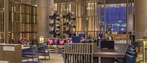 Un moderno vestíbulo de hotel con una zona de estar central, un bar y un amplio ventanal con vistas a la ciudad.