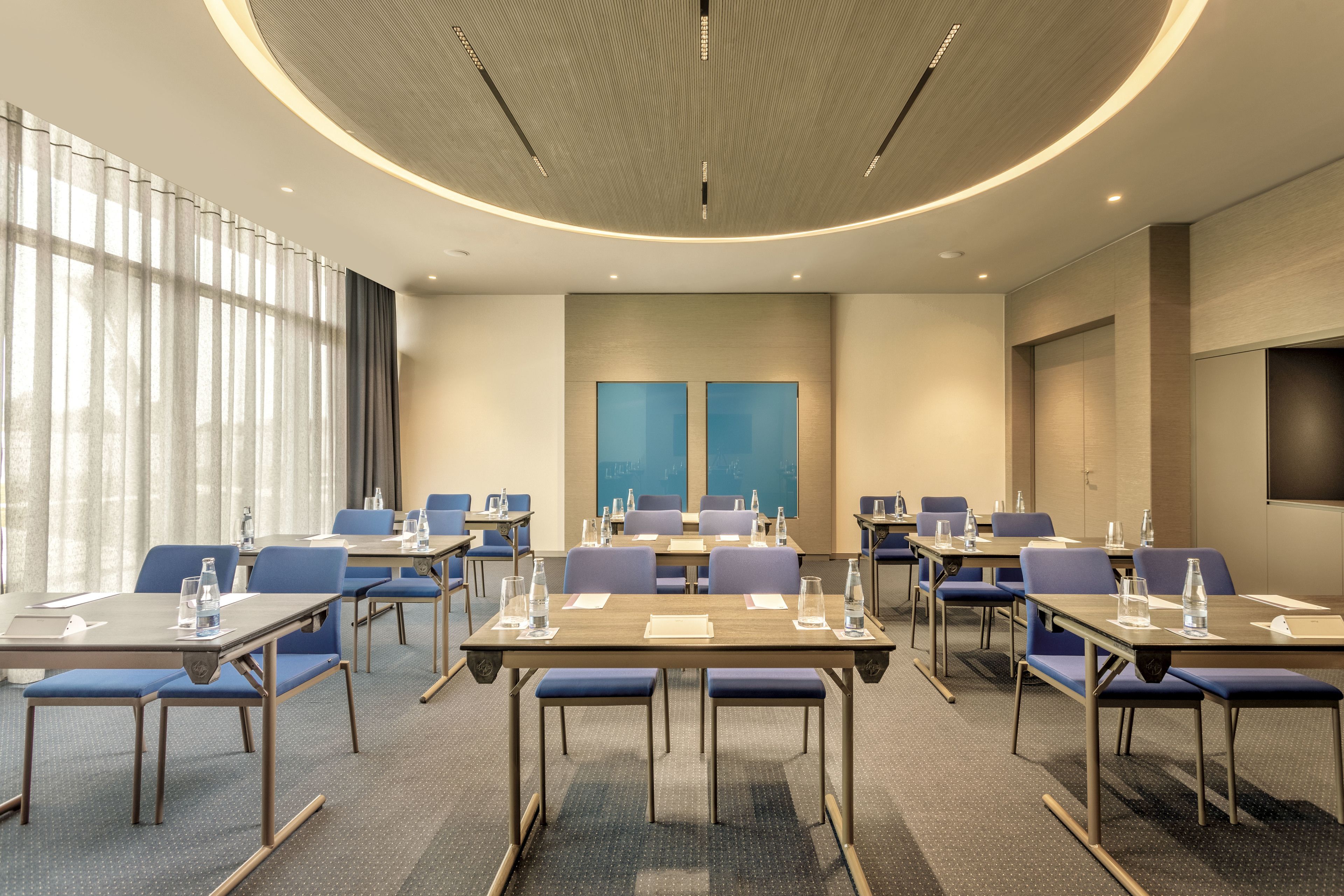 Un salón de conferencias con mesas y sillas, un amplio ventanal con cortinas y un diseño moderno en el techo.