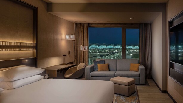 A hotel room with a bed, a sofa, a desk, and a view of a cityscape at night.