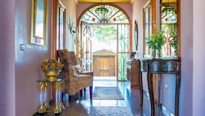 A hallway with a wooden chair, a gold table with a vase, and a staircase leading to a door.