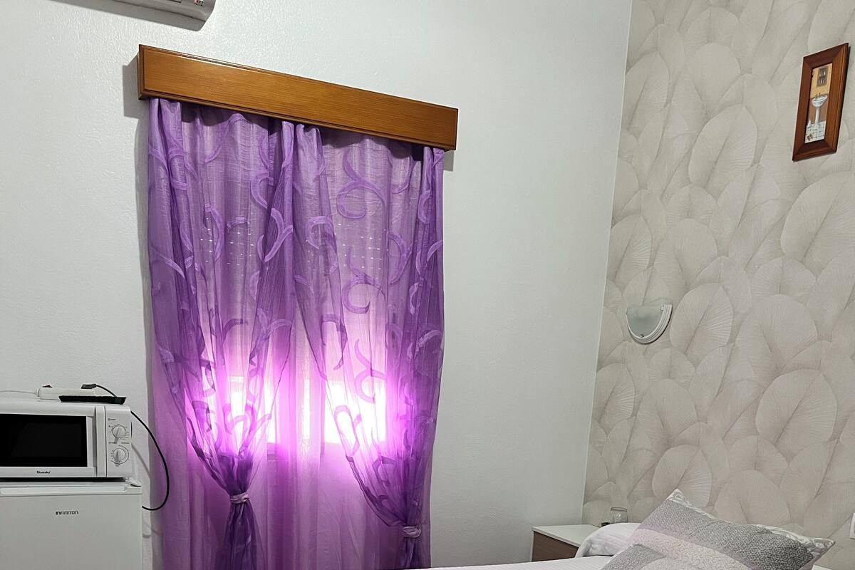 Une chambre à coucher avec un lit, une table de chevet, une chaise, un four à micro-ondes et une fenêtre avec des rideaux violets.