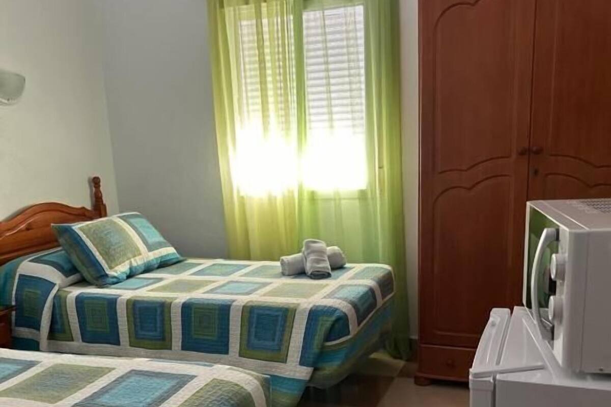 Une chambre avec deux lits, une armoire en bois, un appareil électroménager blanc et une fenêtre avec des rideaux verts.
