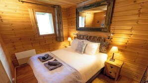 Ein Schlafzimmer in einer Holzhütte mit einem Bett, zwei Nachttischlampen, einem Spiegel und einem Fenster mit Vorhängen.