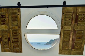 Ein rundes Fenster mit hölzernen Fensterläden, das Meer und Klippen zeigt.