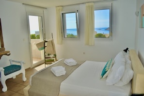 Ein Schlafzimmer mit einem Bett, zwei mit Vorhängen versehenen Fenstern, einem Sessel und Blick aufs Meer.