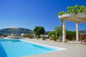 Ein Poolbereich mit Liegestühlen und einer Pergola mit Weinreben, vor einer Bergkulisse.