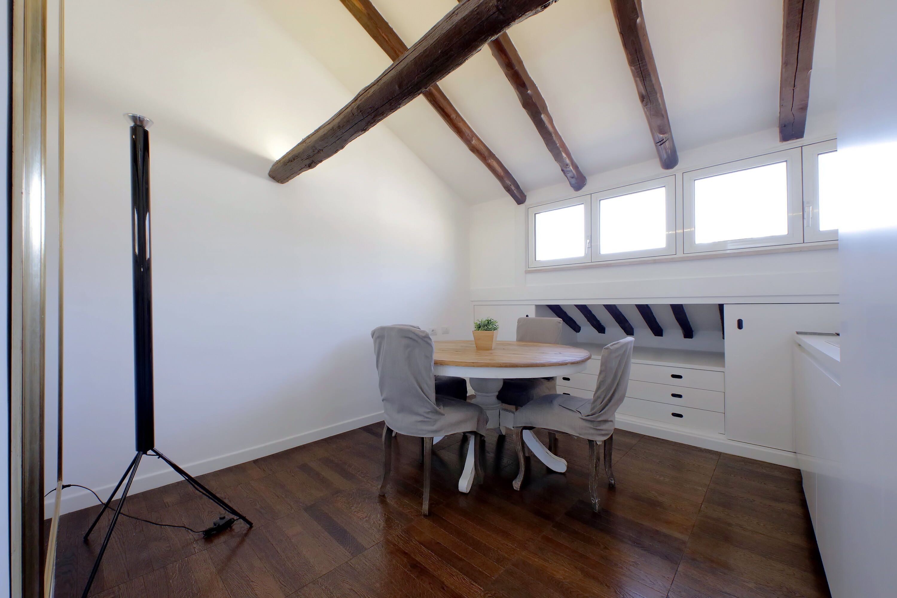 Une pièce avec un plancher en bois, une table ronde entourée de chaises, un trépied et une fenêtre avec des stores.