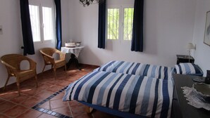 Een kamer met een bed, twee rieten stoelen, een klein tafeltje met een mand en een kroonluchter.