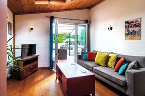 A cozy living room with a wooden ceiling, a television on a wooden stand, a grey sofa with colorful cushions, a wooden coffee table, and a glass door leading outside. - Charming cottage with large pool, garden & views of Pyrénées, near Carcassonne (Montazels)
