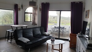 Un salon moderne avec un canapé en cuir noir, une petite table ronde ornée d’un vase, une table à manger avec des chaises et un balcon offrant une vue sur la verdure.