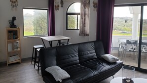 Un salon moderne avec un canapé en cuir noir, une table basse en bois et une table à manger. Il y a deux grandes fenêtres avec des rideaux violets, un attrape-rêves et une suspension.