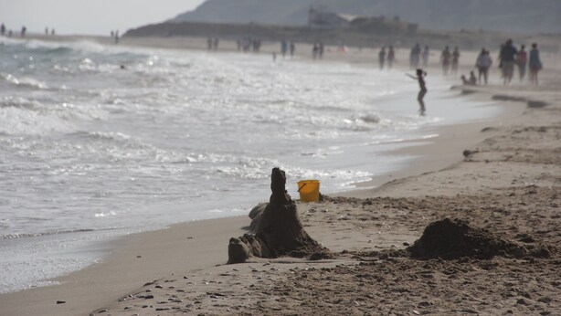 Une plage avec des personnes au loin, un château de sable et un seau jaune.
