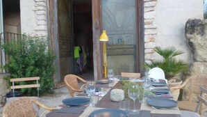 Un espace extérieur aménagé pour les repas, avec des chaises en osier et une table dressée, agrémenté d’un mur en pierre et d’une porte en bois.