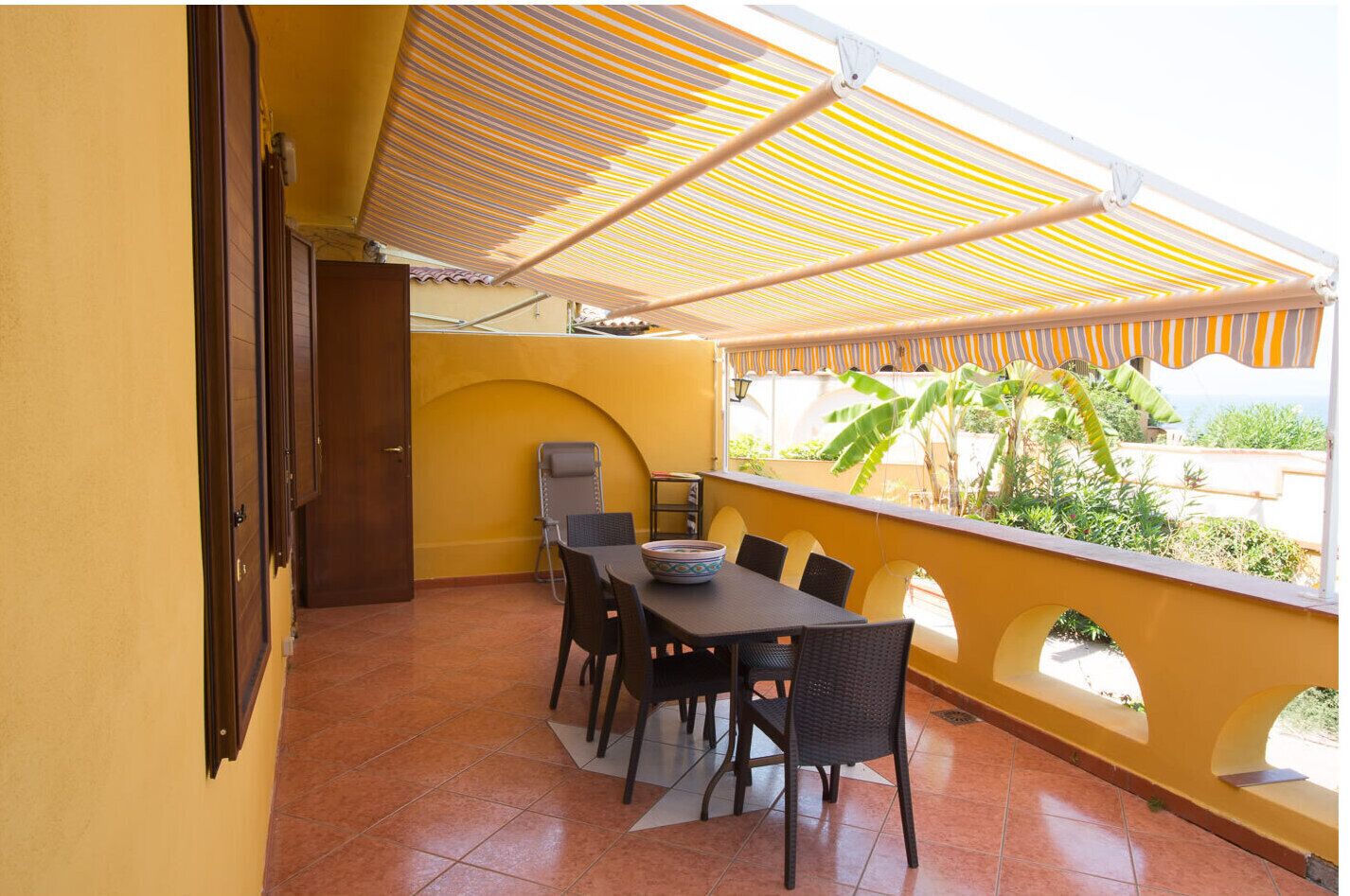 Un balcon avec une table et des chaises, un fauteuil, une porte et une vue sur la verdure et les bâtiments.