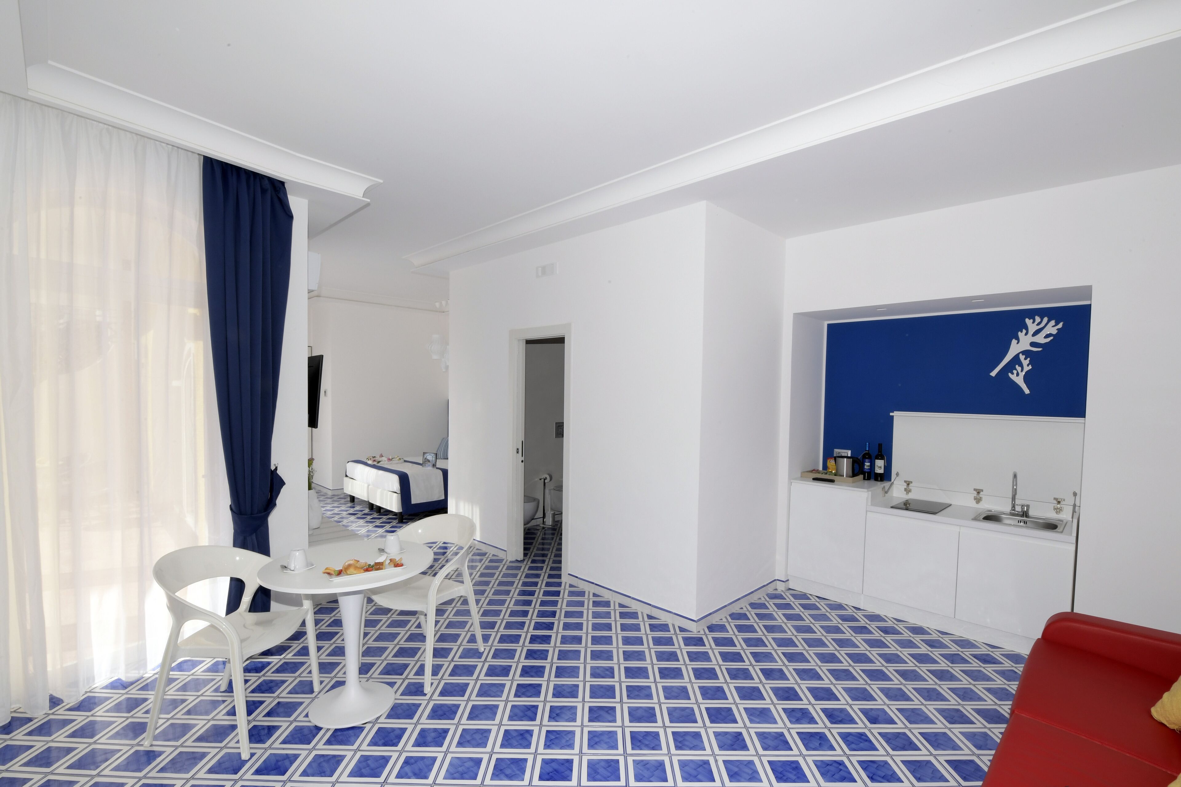 A modern hotel room with a blue-tiled floor, white furniture, a red sofa, and a kitchen area with a blue backsplash.
