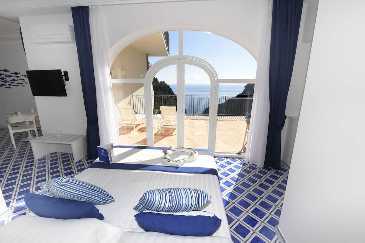 A hotel room with a bed, a small table, and a chair. There is a balcony with a view of the sea.