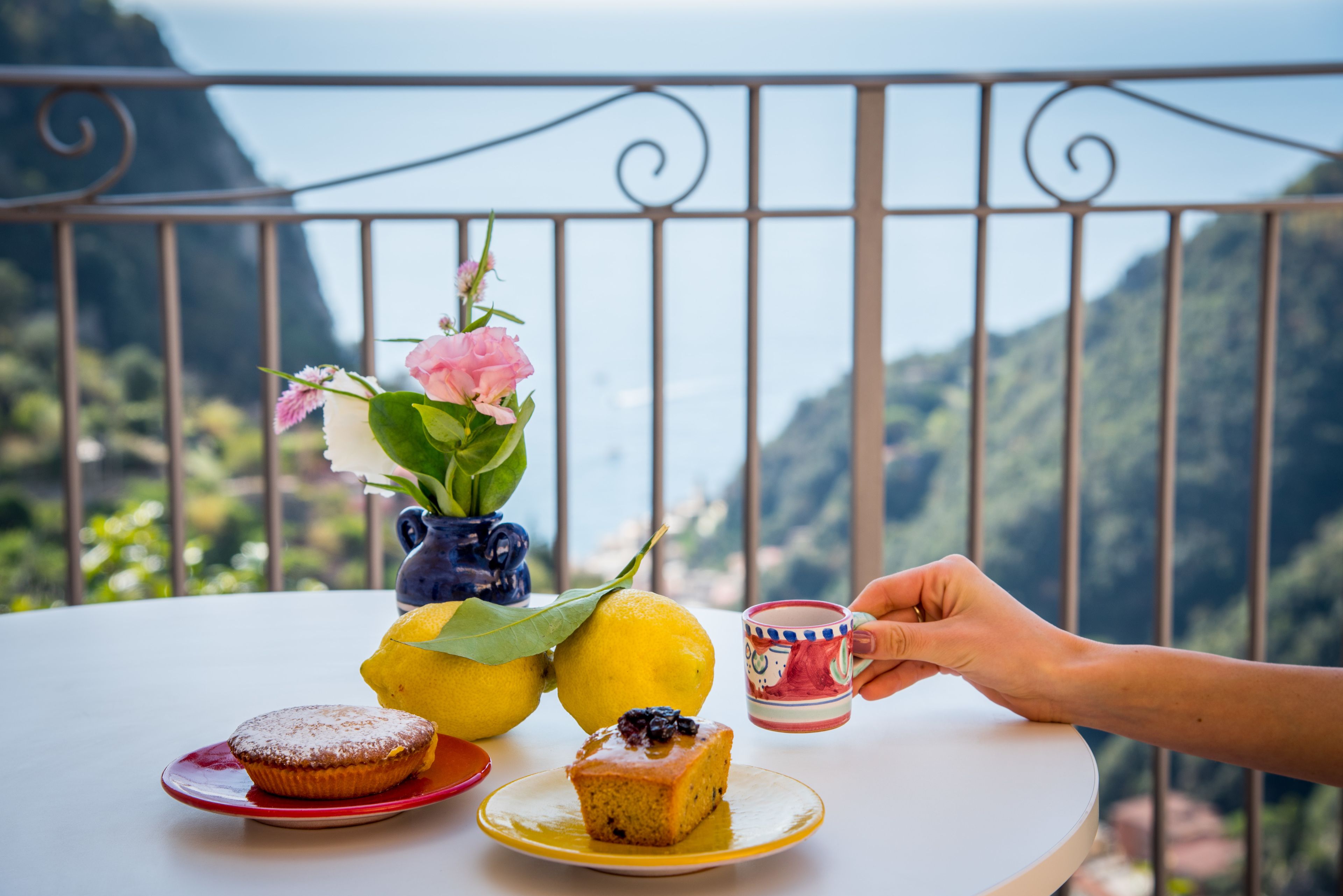 Una mesa con un postre, un florero con flores y una taza, frente a un paisaje montañoso.