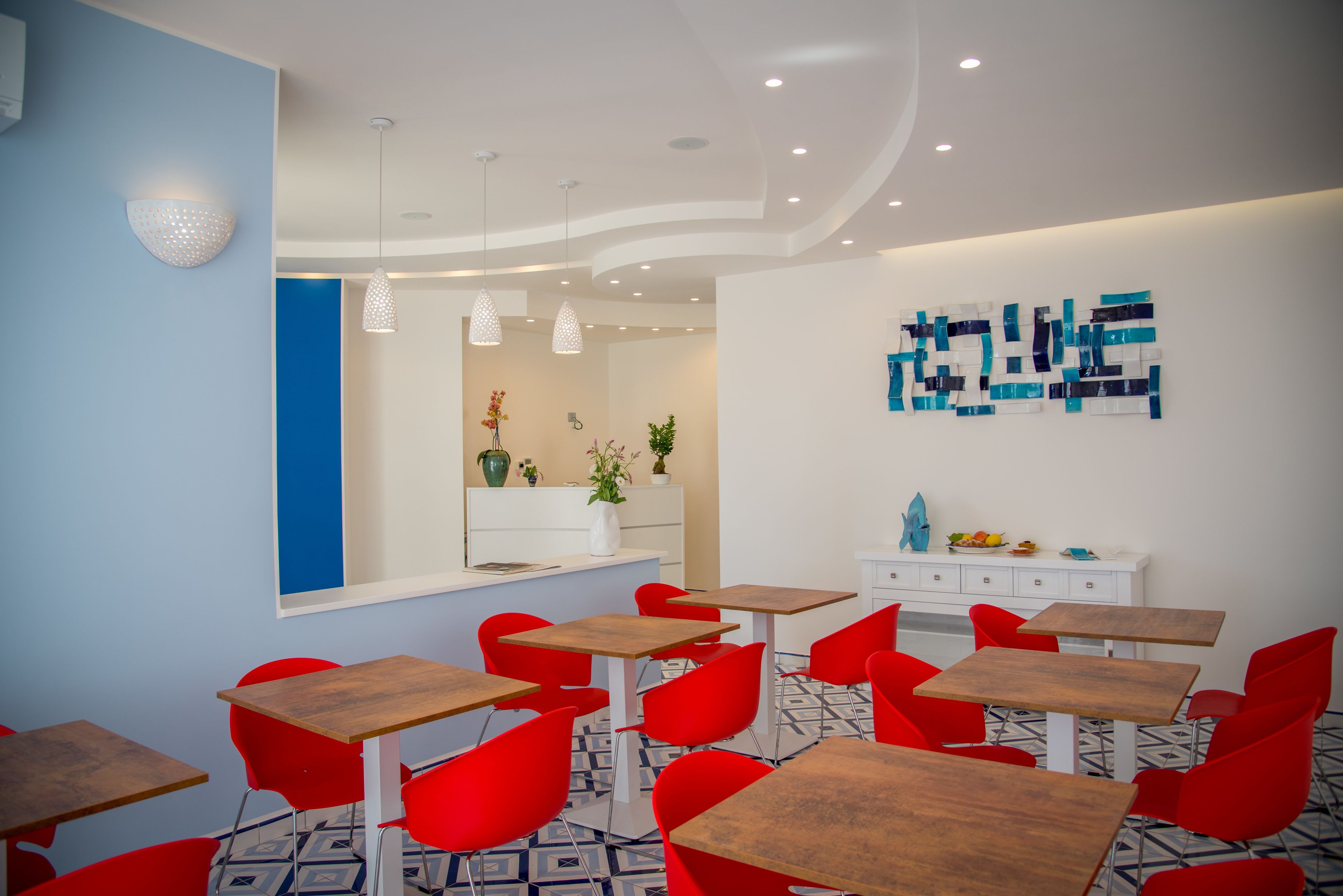 Un espace repas moderne avec des chaises rouges et des tables en bois, un comptoir blanc agrémenté d’un vase et de plantes, et une œuvre d’art murale aux couleurs bleue et blanche.