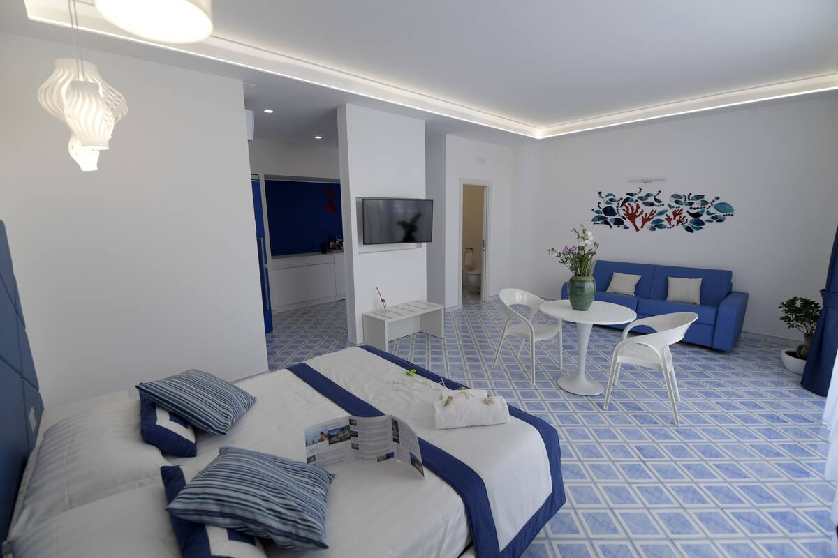 A modern hotel room with a large bed, a TV, a small dining area, and a blue sofa.