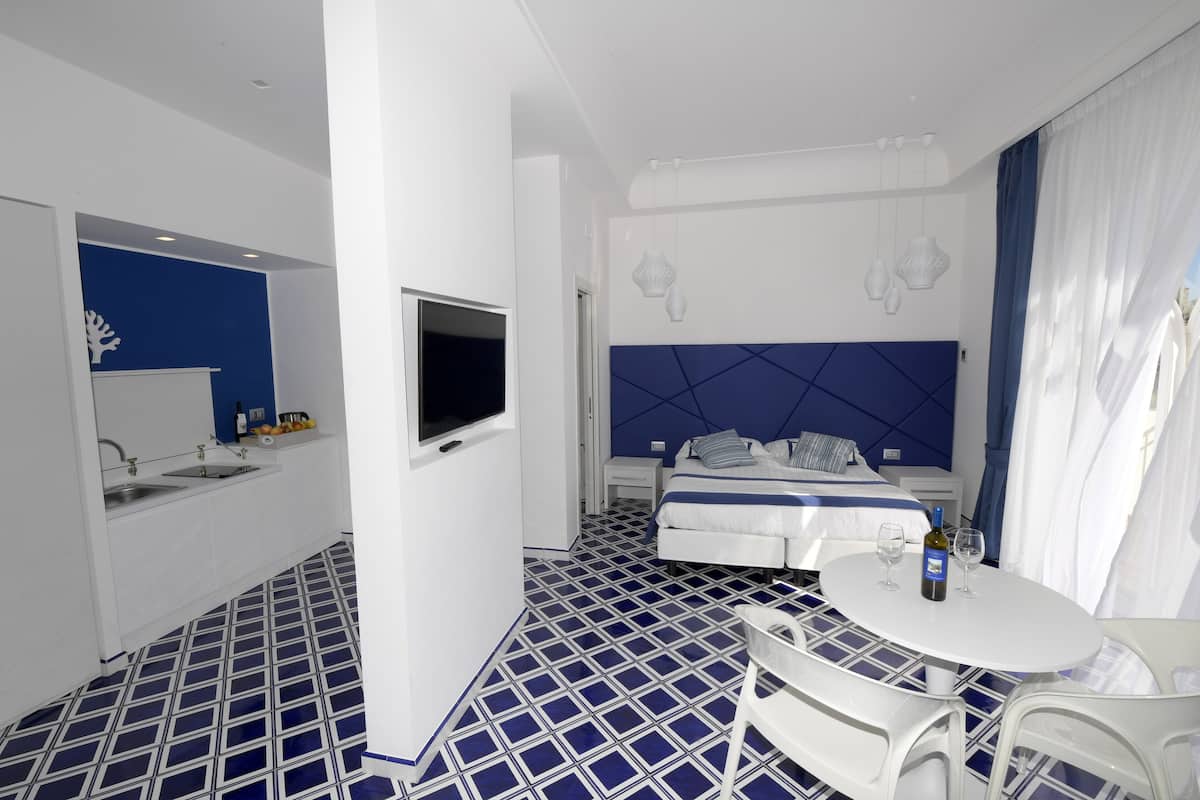A hotel room with a bed, a TV, a small table with a bottle and glasses, a sink, and a blue and white tiled floor.
