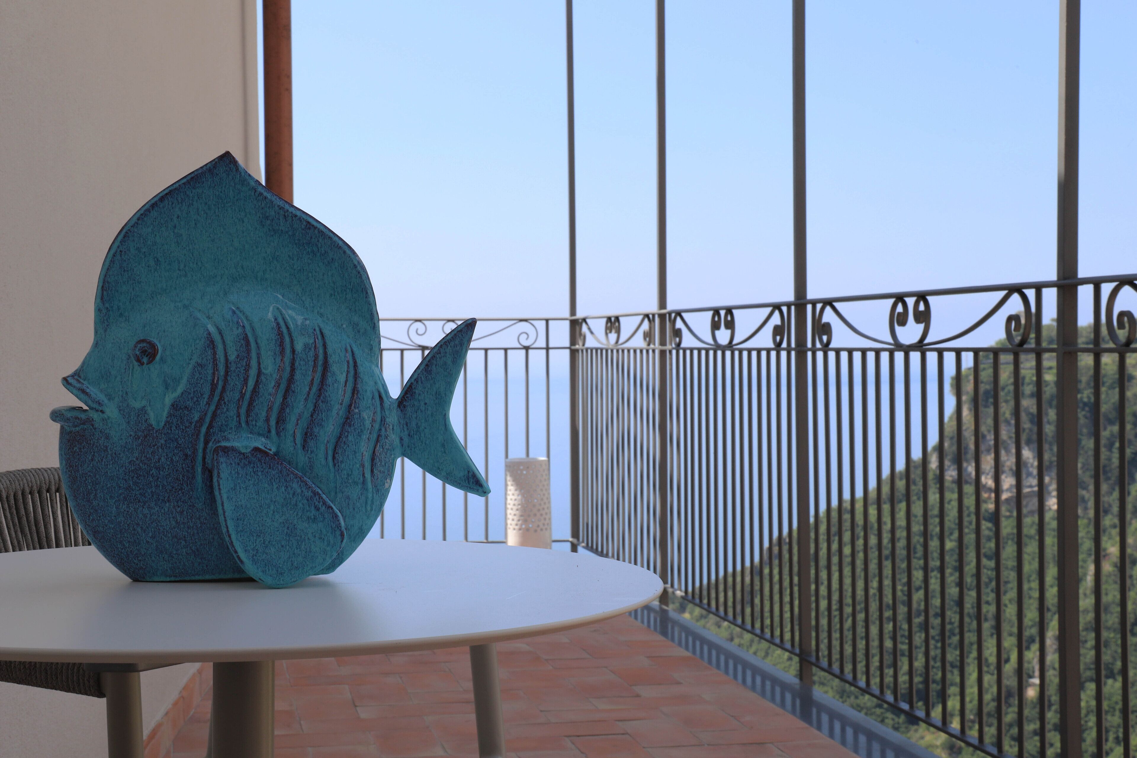 Een blauwe sculptuur van een vis op een tafel, met uitzicht op een bergachtig landschap door een balkonhek.