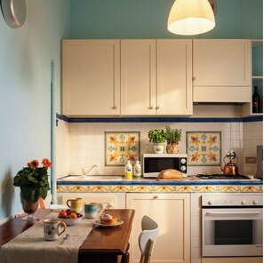 A kitchen with cabinets, a microwave, a kettle, a table with a chair, and a vase with flowers.