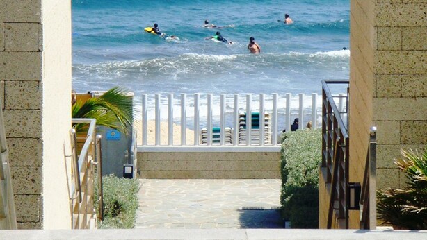 Un escalier en pierre mène à une plage où l’on peut voir des gens nager et faire de la planche à voile.