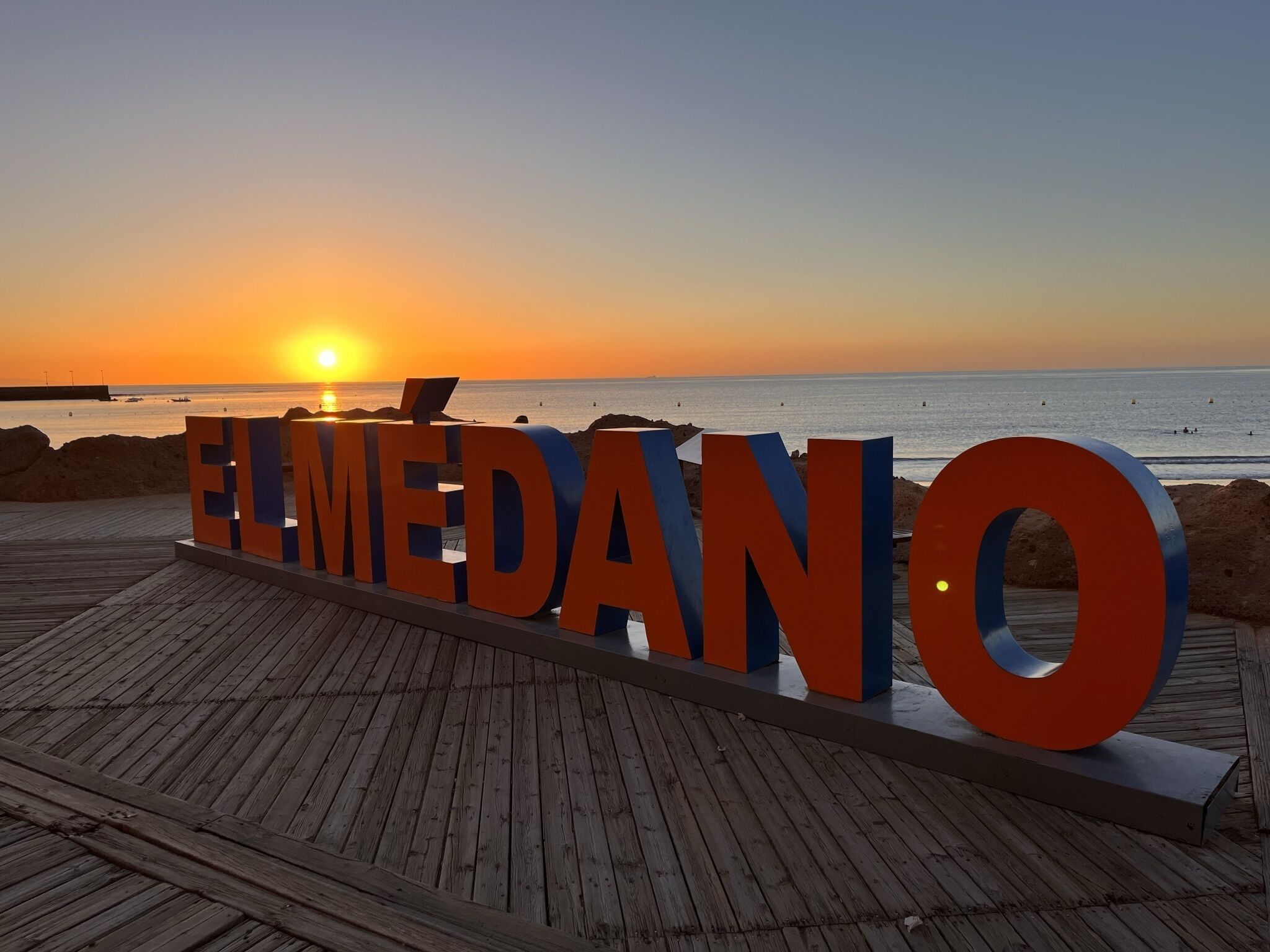 Un muelle de madera con letras grandes y llamativas que deletrean "EL MEDANO" sobre un fondo de atardecer.
