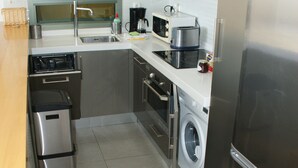 Une cuisine compacte équipée d’appareils électroménagers en acier inoxydable, d’un lave-linge et d’un tabouret en bois.