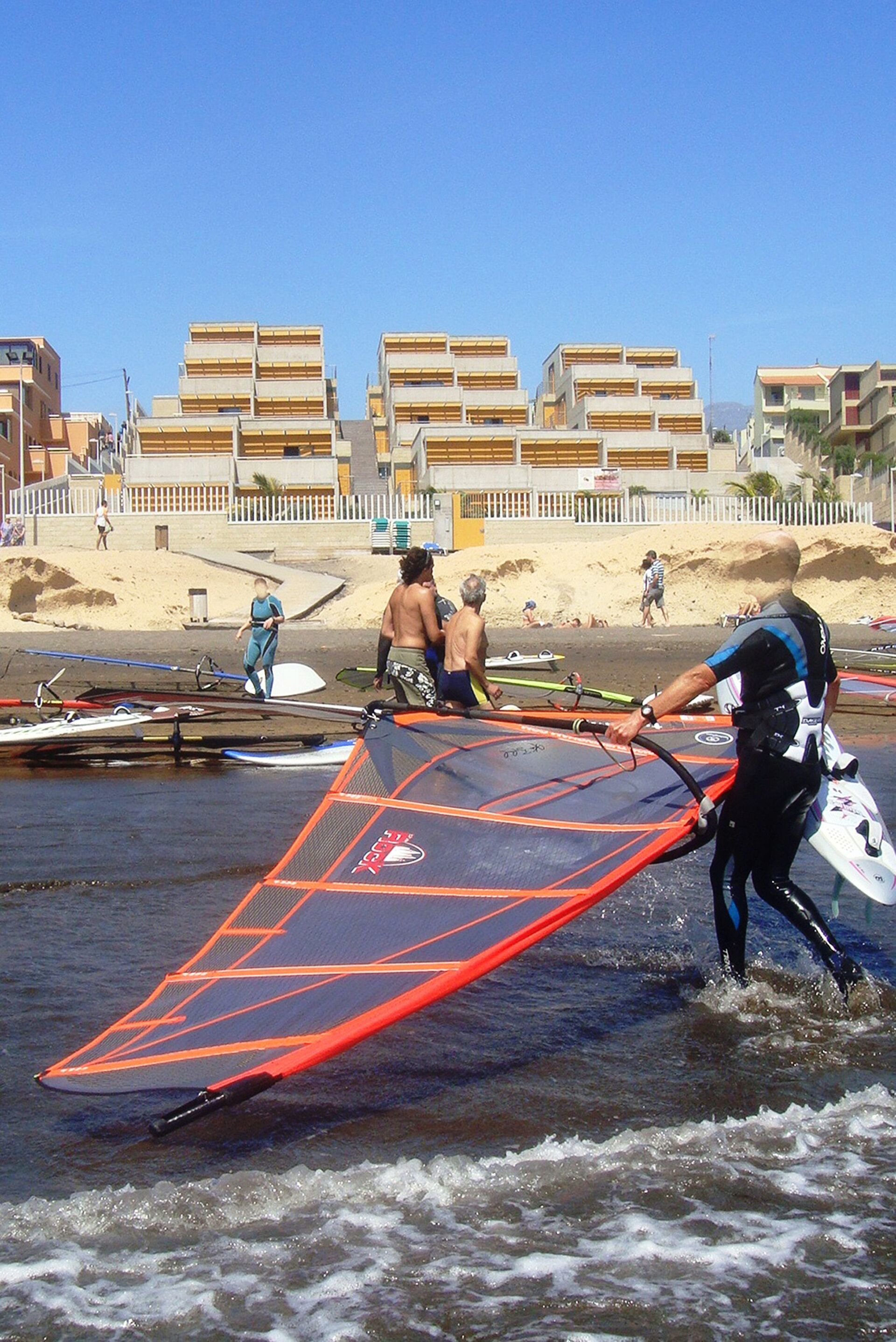 Una playa con gente y tablas de surf, cielo despejado y edificios al fondo.