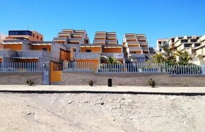 A row of modern, multi-story residential buildings with balconies, a paved area in front, and a clear blue sky. - Vacation home on the beach, 2 bedrooms, 30 m² terrace. Beach 30 seconds (El Médano, Granadilla, Tenerife)