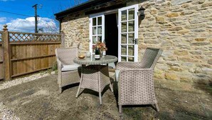 A stone building with a wooden fence, two wicker chairs, and a table with a vase and a bottle. - The Brambles - one bedroom Country Cottage near Dorset Coast. (Weymouth)