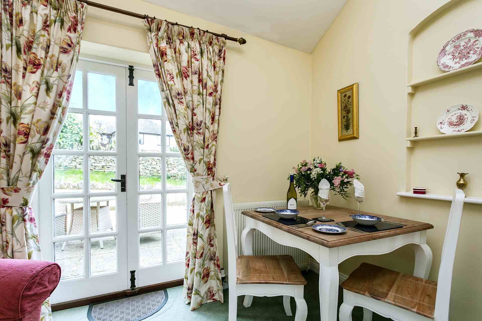Een eetruimte met een tafel voor twee, een stoel, een bloemen gordijn en uitzicht door een glazen deur.