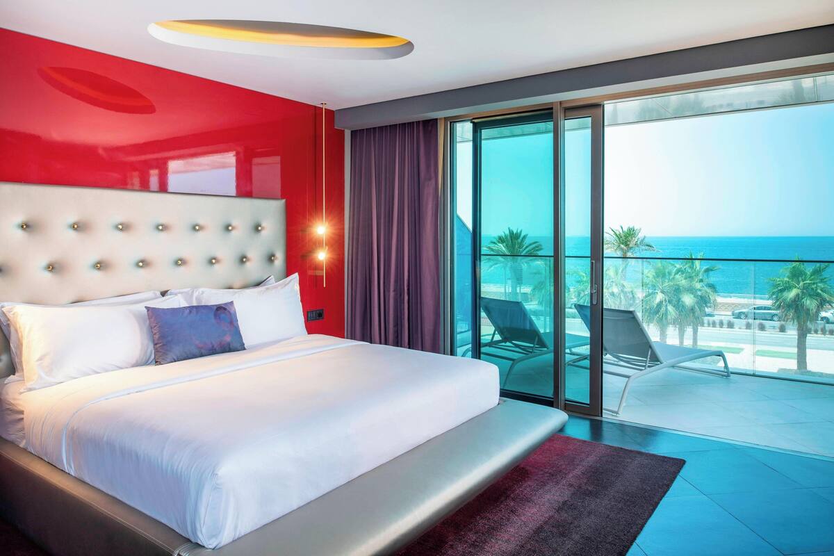 Een moderne hotelkamer met een groot bed, een rode accentmuur en een balkon met uitzicht op de oceaan.