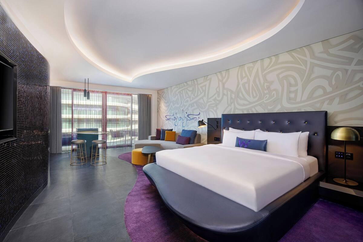 Een moderne hotelkamer met een groot bed, een zithoek met krukjes, een televisie en een wand met abstracte patronen.