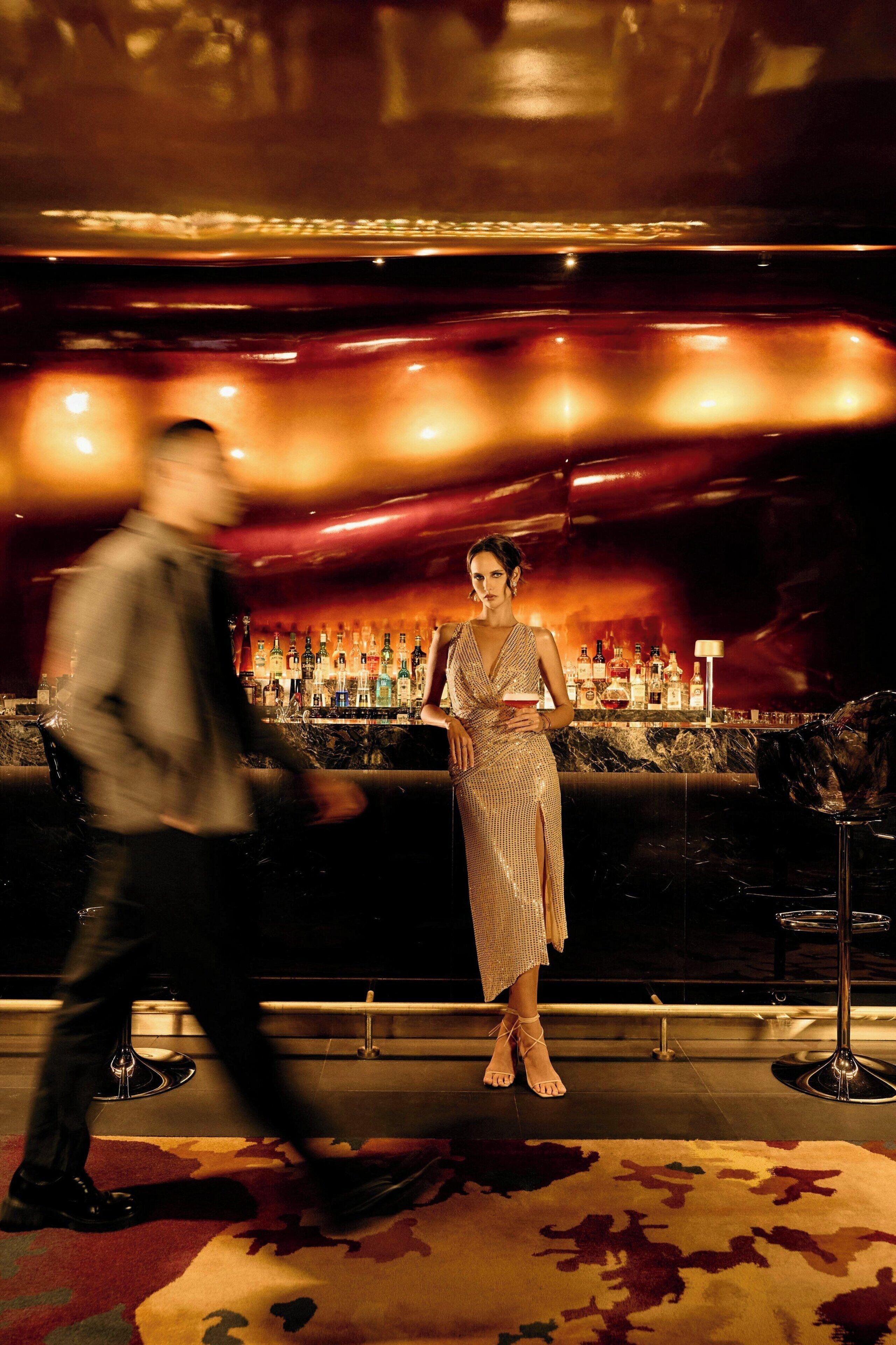 A bar area with a woman standing, a man walking, and a bar counter with various bottles.