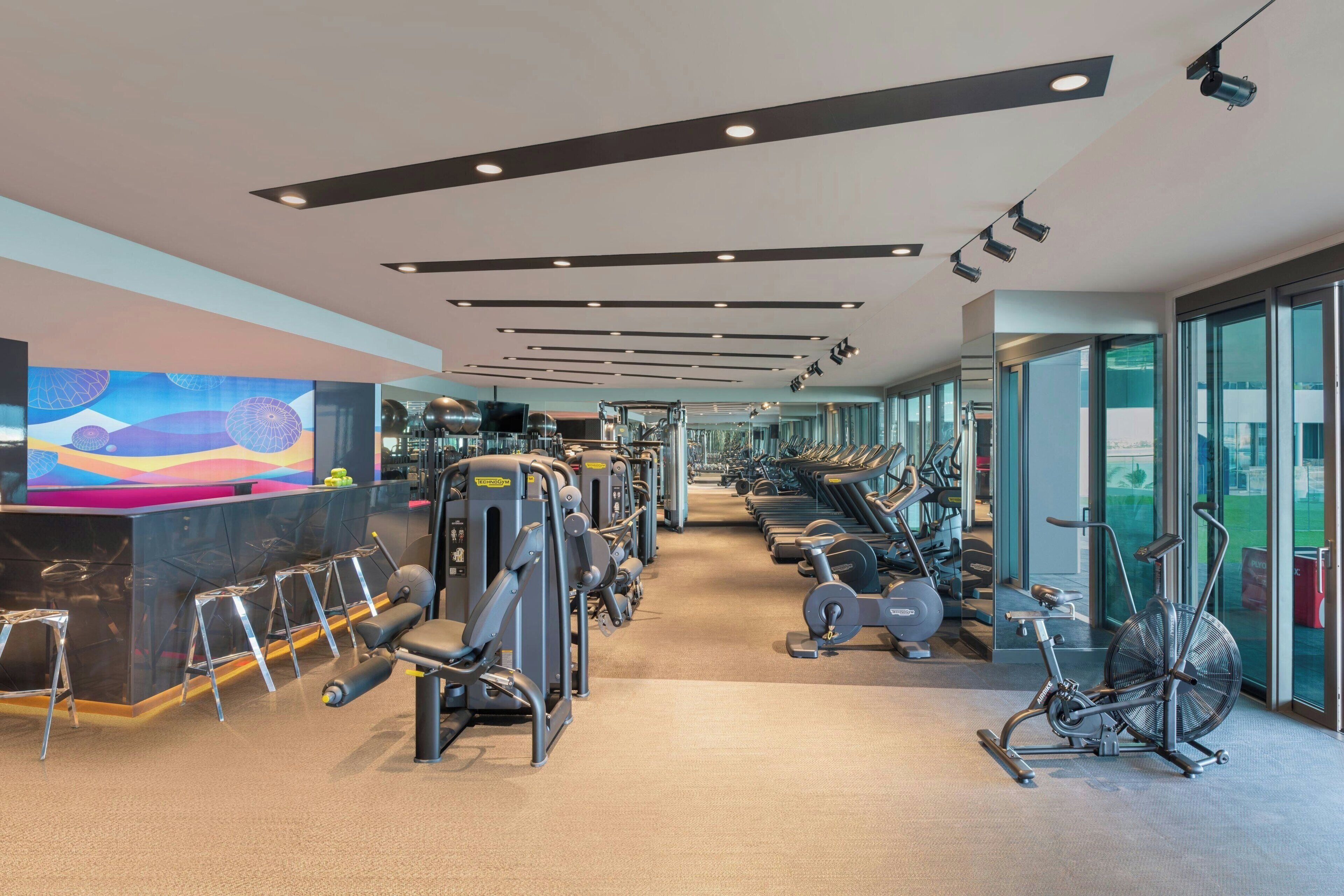 Un gimnasio moderno con diversos equipos de ejercicio, una zona de recepción y amplias ventanas con vistas.