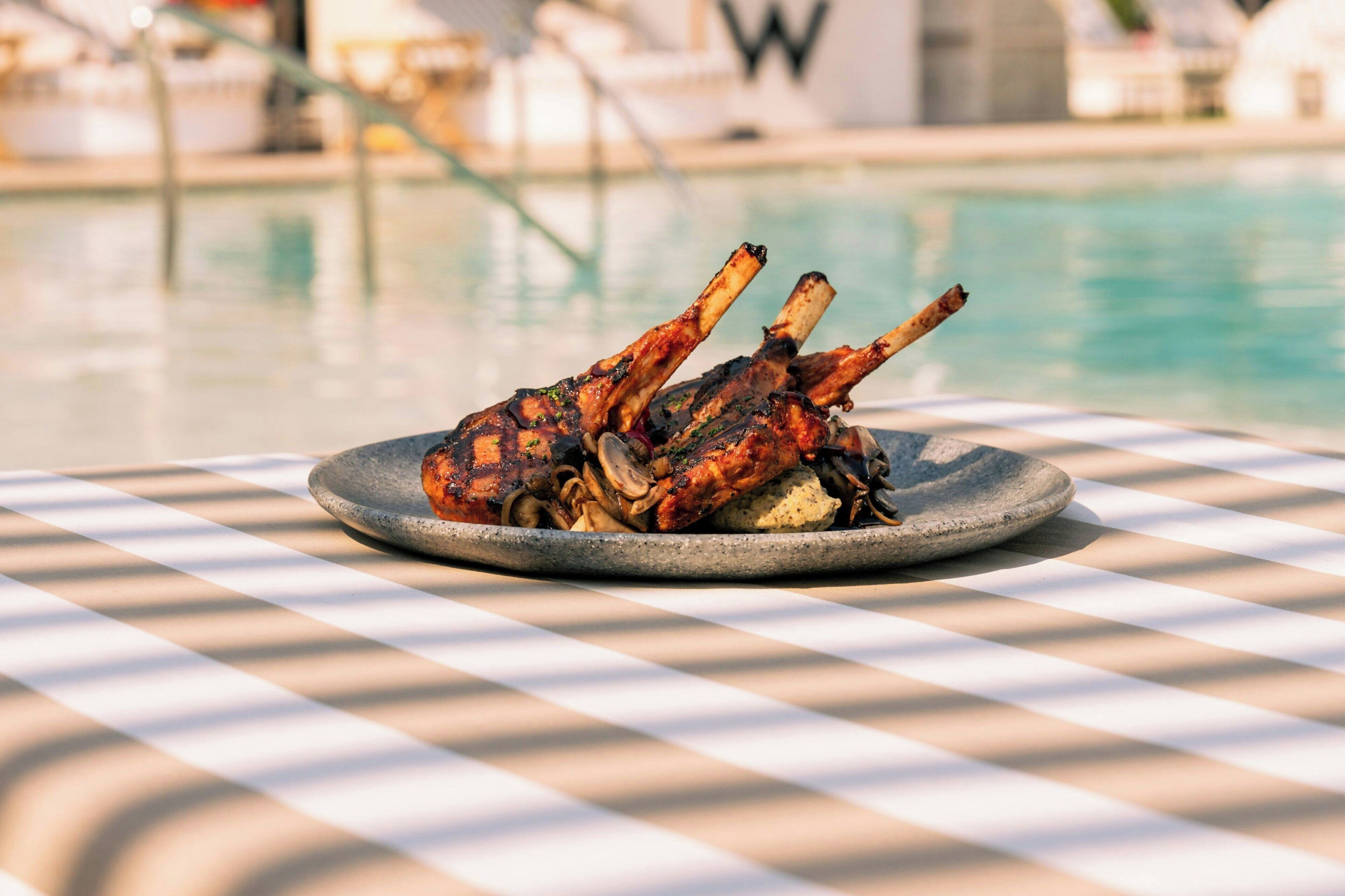 A table with a checkered tablecloth, a plate of grilled lamb chops, and a pool in the background.