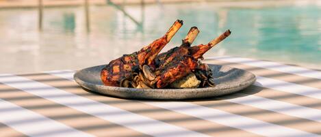 A table with a checkered tablecloth, a plate of grilled lamb chops, and a pool in the background.