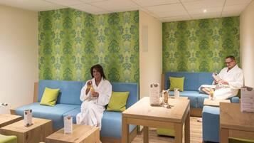 A spa room with two blue sofas, wooden tables, and green patterned wallpaper.
