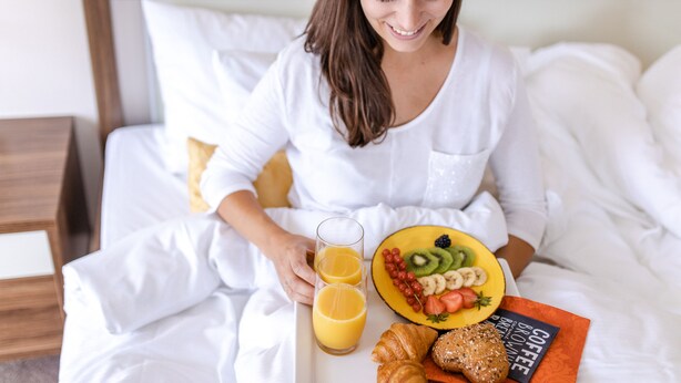 Une personne est assise sur un lit, un plateau de petit-déjeuner devant elle, contenant une assiette de fruits, des croissants et un livre sur le café.