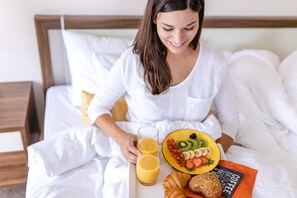 Une personne est assise sur un lit, un plateau de petit-déjeuner devant elle, contenant une assiette de fruits, des croissants et un livre sur le café.
