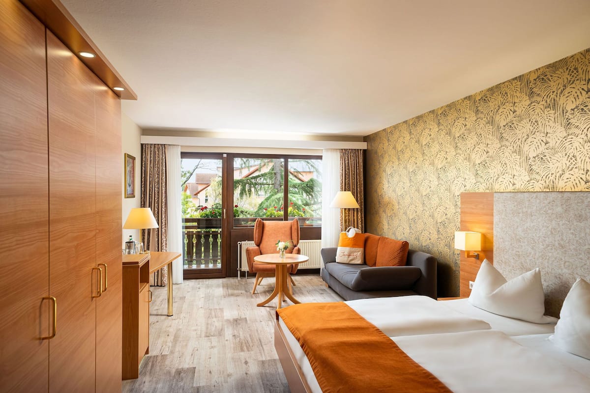 Ein Hotelzimmer mit einem Bett, einem Sofa, einem kleinen Tisch und einem Sessel. Es gibt einen Balkon mit Blick auf Grünflächen und Gebäude.