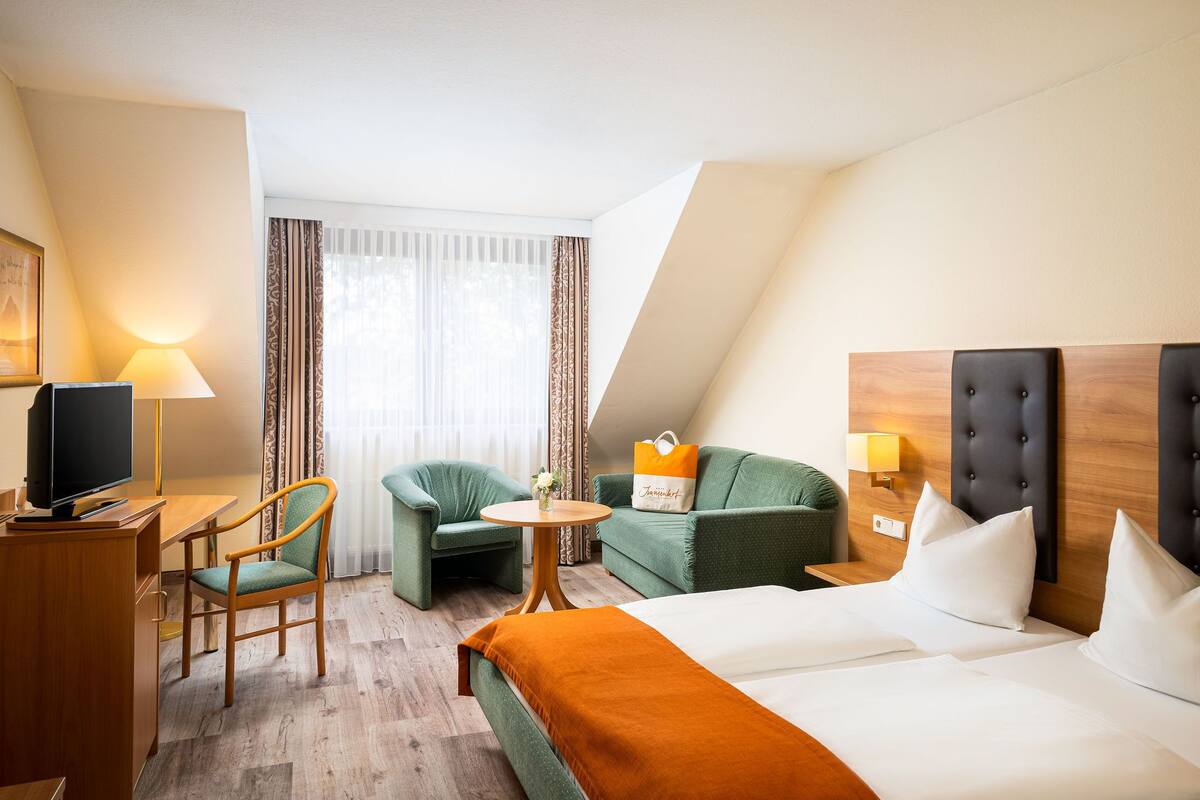 Ein Hotelzimmer mit einem Bett, einem Schreibtisch mit Fernseher, zwei Sesseln, einem kleinen runden Tisch mit Blumenvase und einem Sofa mit Kissen.