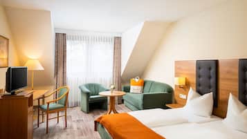 A hotel room with a bed, a desk with a television, two armchairs, a small round table with a flower vase, and a sofa with a pillow.