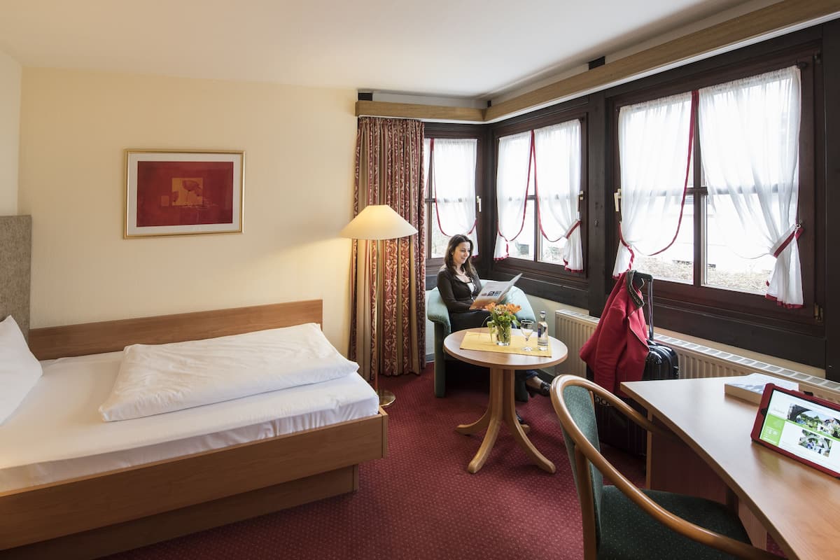 Ein Hotelzimmer mit einem Bett, einem Tisch mit Lampe, einem Stuhl, einem Fenster mit Vorhängen und einem gerahmten Bild an der Wand.