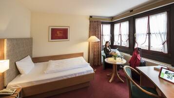 A hotel room with a bed, a table with a lamp, a chair, a window with curtains, and a framed picture on the wall.